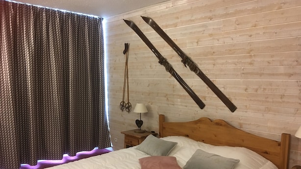 A bedroom with a wooden headboard, a bed with white bedding and multiple pillows, a nightstand with a lamp, and a window with curtains.