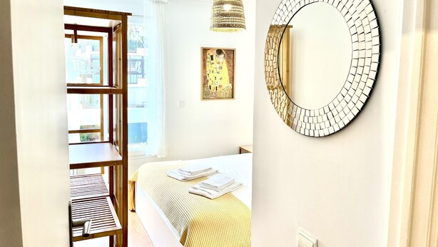 A bedroom with a bed, a wooden shelf, a hanging light, a wall-mounted mirror, and a painting. - Guia Acacias Apartment (Cascais)