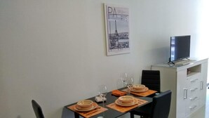 Un coin repas avec une table dressée pour deux personnes, deux chaises, un téléviseur posé sur un meuble et un tableau encadré représentant Paris accroché au mur.