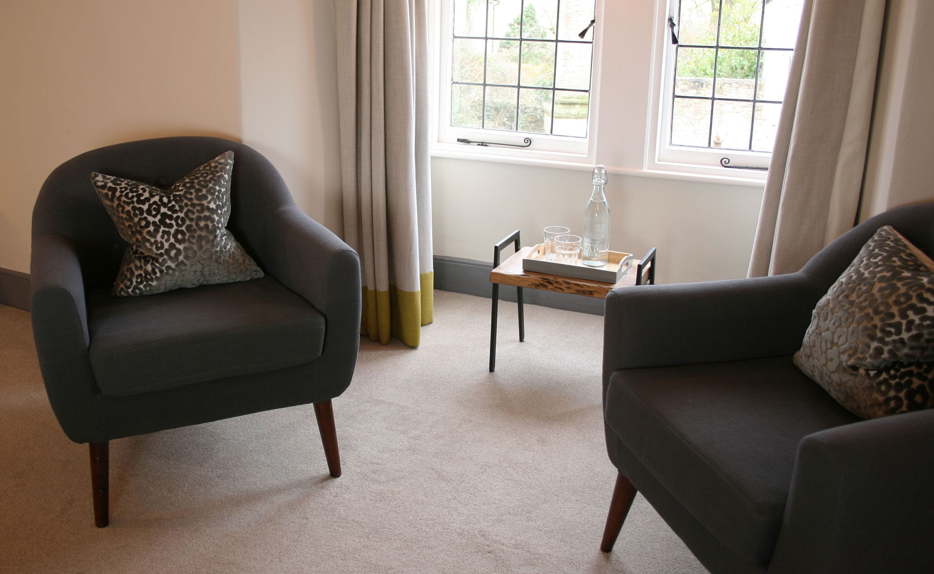 A room with two dark armchairs, one with a leopard print pillow, and a small wooden table with a bottle and glasses.