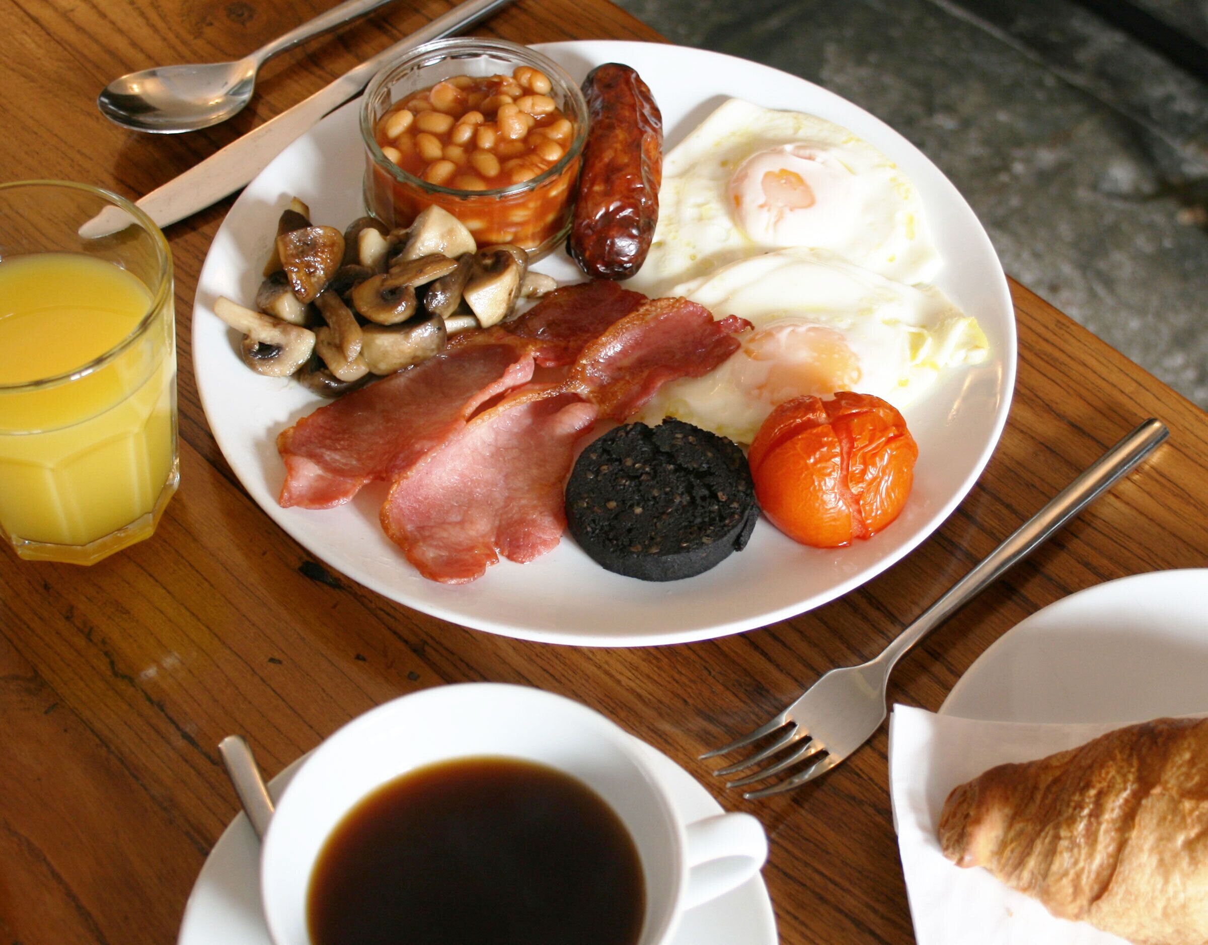 Un plato de desayuno con huevos, salchichas, frijoles, champiñones, tocino y morcilla, además de un vaso de jugo de naranja y una taza de café.