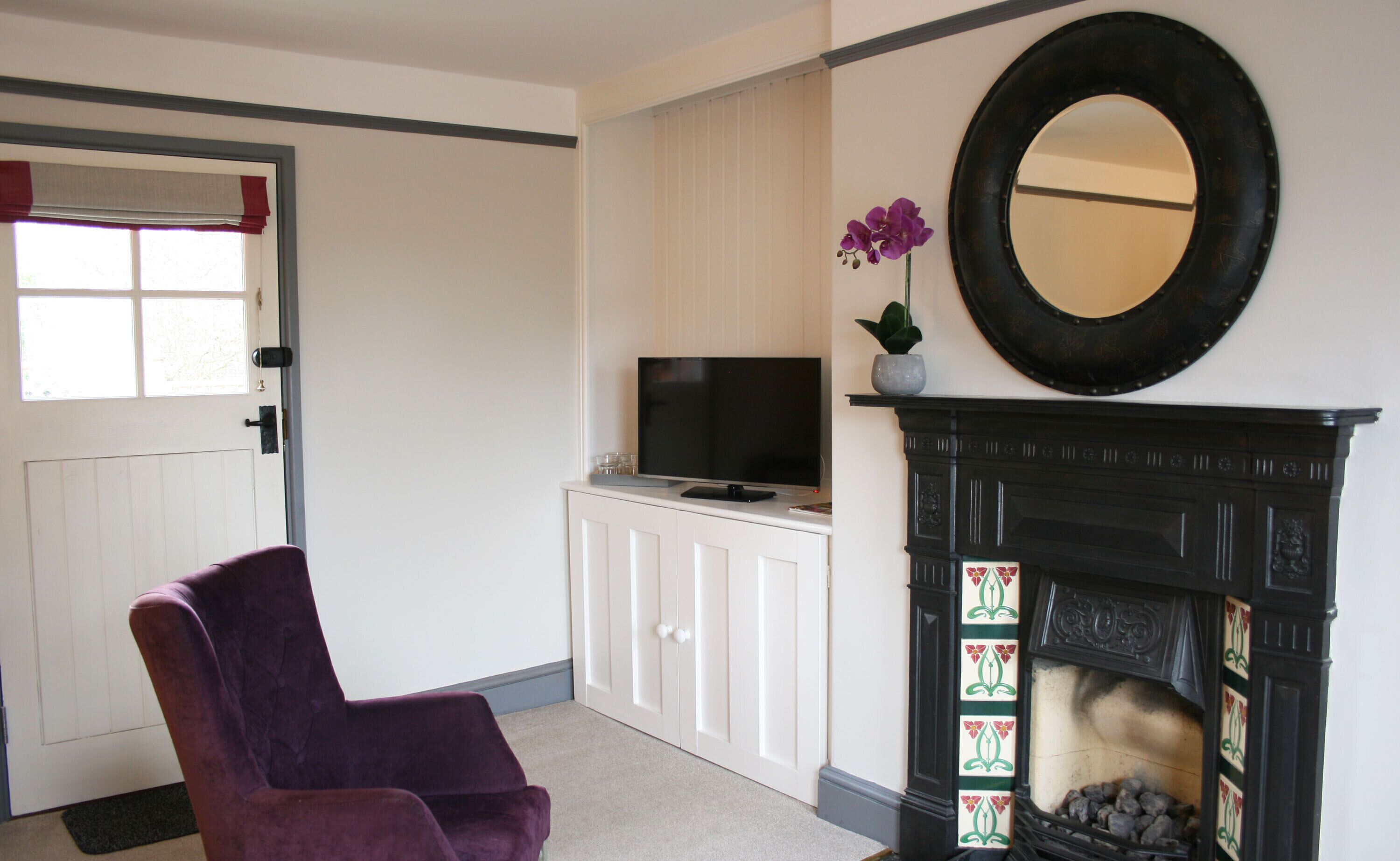 Een woonkamer met een paarse fauteuil, een televisie op een kast, een open haard en een spiegel aan de muur.