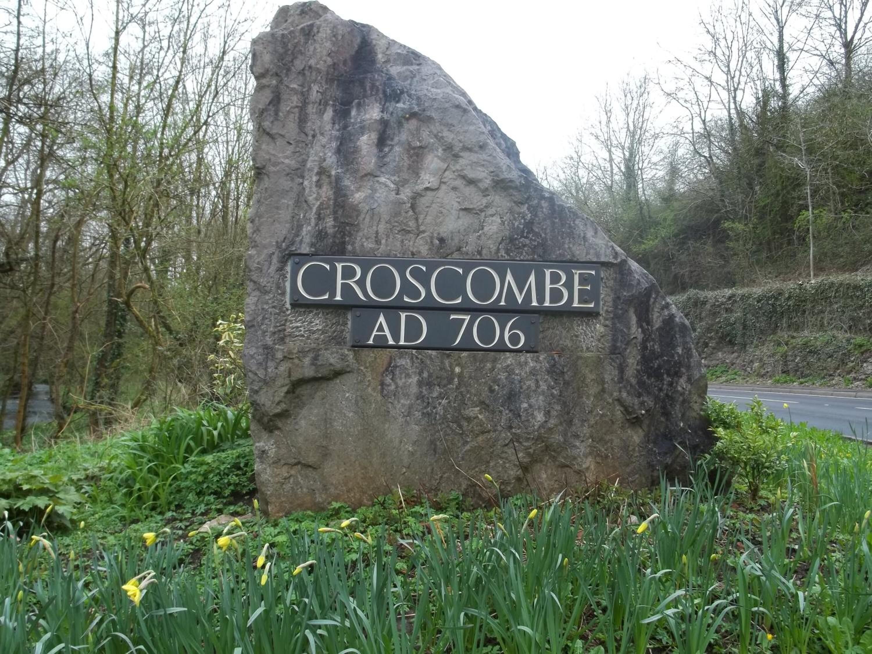 En stor sten med en skylt som visar texten ”CROSCOMBE AD 706”, omgiven av grönska och påskliljor.