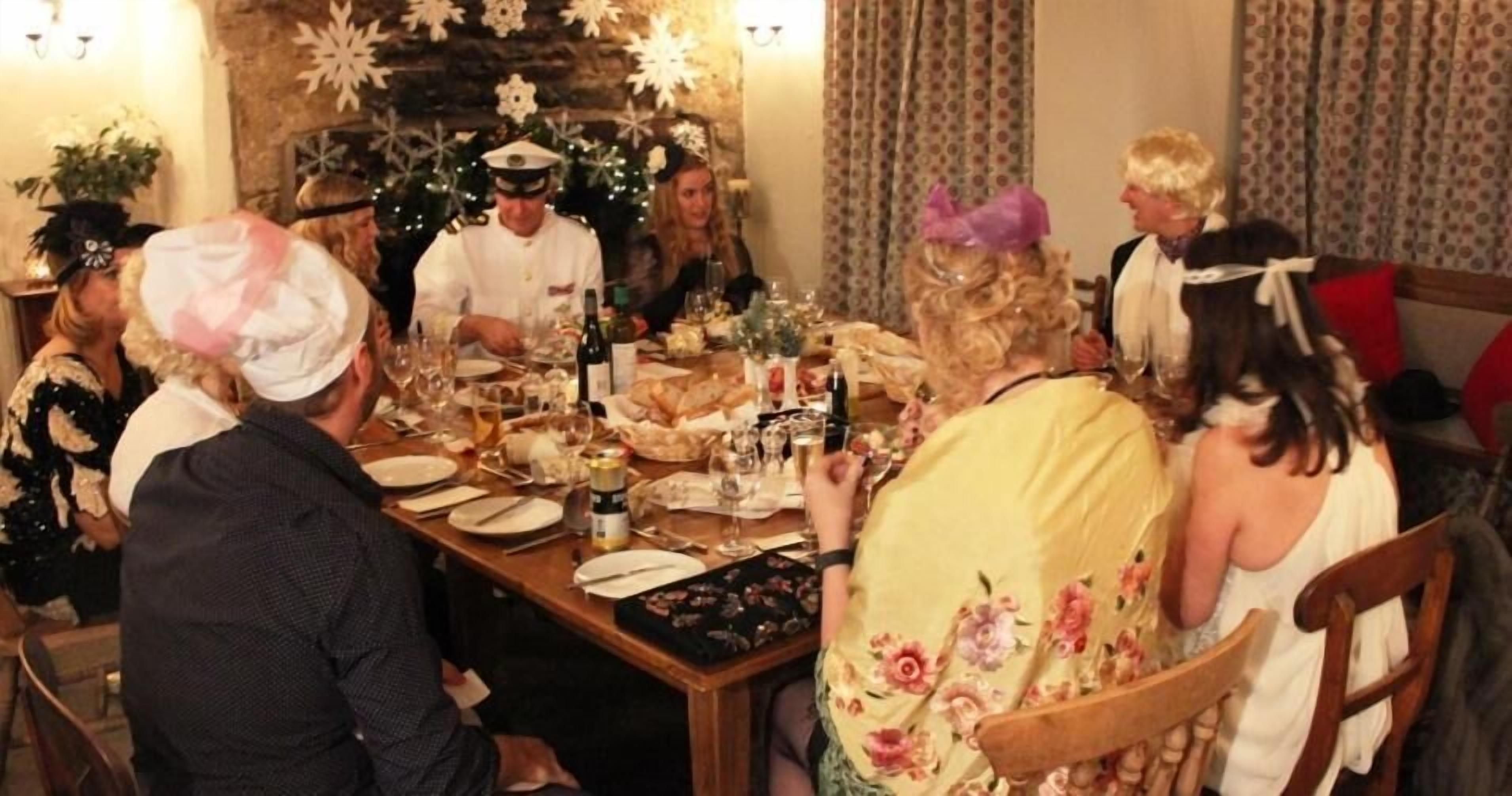 Een groep mensen zit rond een houten eettafel, met een kok in uniform, feestelijke decoraties en een kerstboom op de achtergrond.
