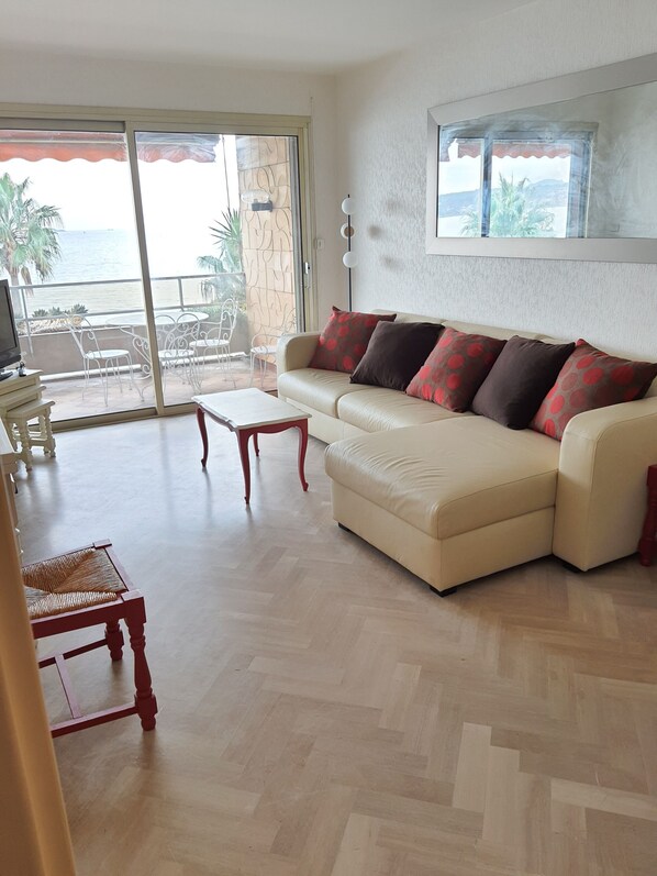 A living room with a beige sofa, a wooden coffee table, and a television. There is a sliding glass door leading to an outdoor area with chairs and a view of the ocean.