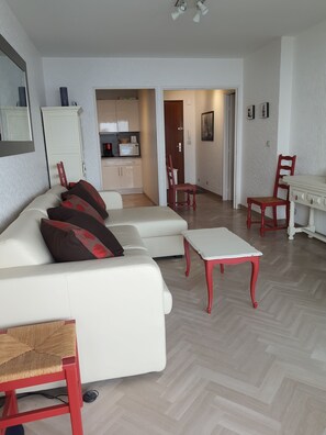 Ein modernes Wohnzimmer mit einem weißen Sofa, roten Sesseln und einem Esstisch. Im Hintergrund ist eine Küche zu sehen.