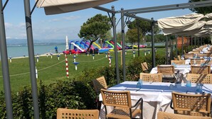 An outdoor dining area with tables set for guests, overlooking a scenic view of a lake and beach. - Lakefront apartment with pool and WiFi (Sirmione)