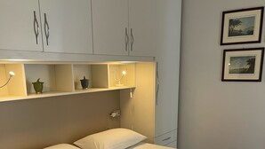 A neatly made bed with white linens and a beige blanket, a bedside table with a lamp, and a shelf with decorative items. - Lakefront apartment with pool and WiFi (Sirmione)