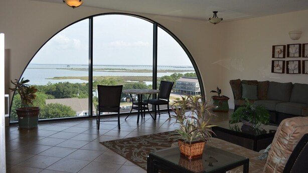 A spacious living area with a large arched window offering a view of the water and a bridge. There is a sofa, a coffee table, and several chairs. - Best seat in the house! FALL RATES, NO CROWDS, RENT A GOLF CART, WALK THE BEACH. (Dauphin Island)