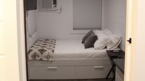Un dormitorio compacto con cama individual, mesita de noche pequeña, ventana con persianas, aire acondicionado de pared y suelo de madera.