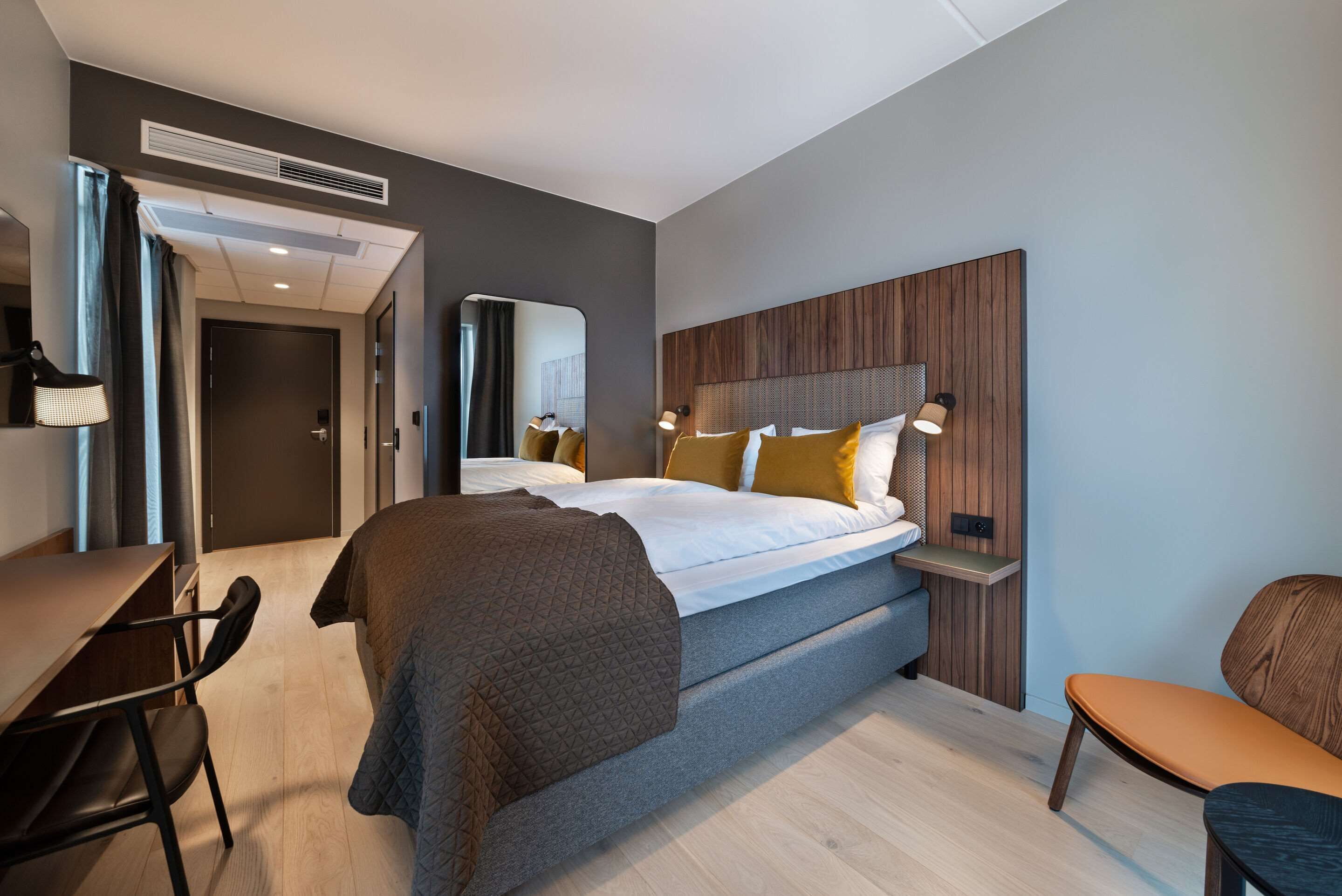 A modern hotel room with a bed, bedside table, lamp, and a desk with a chair.