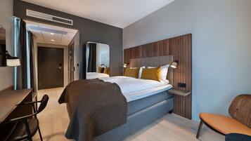A modern hotel room with a bed, bedside table, lamp, and a desk with a chair.