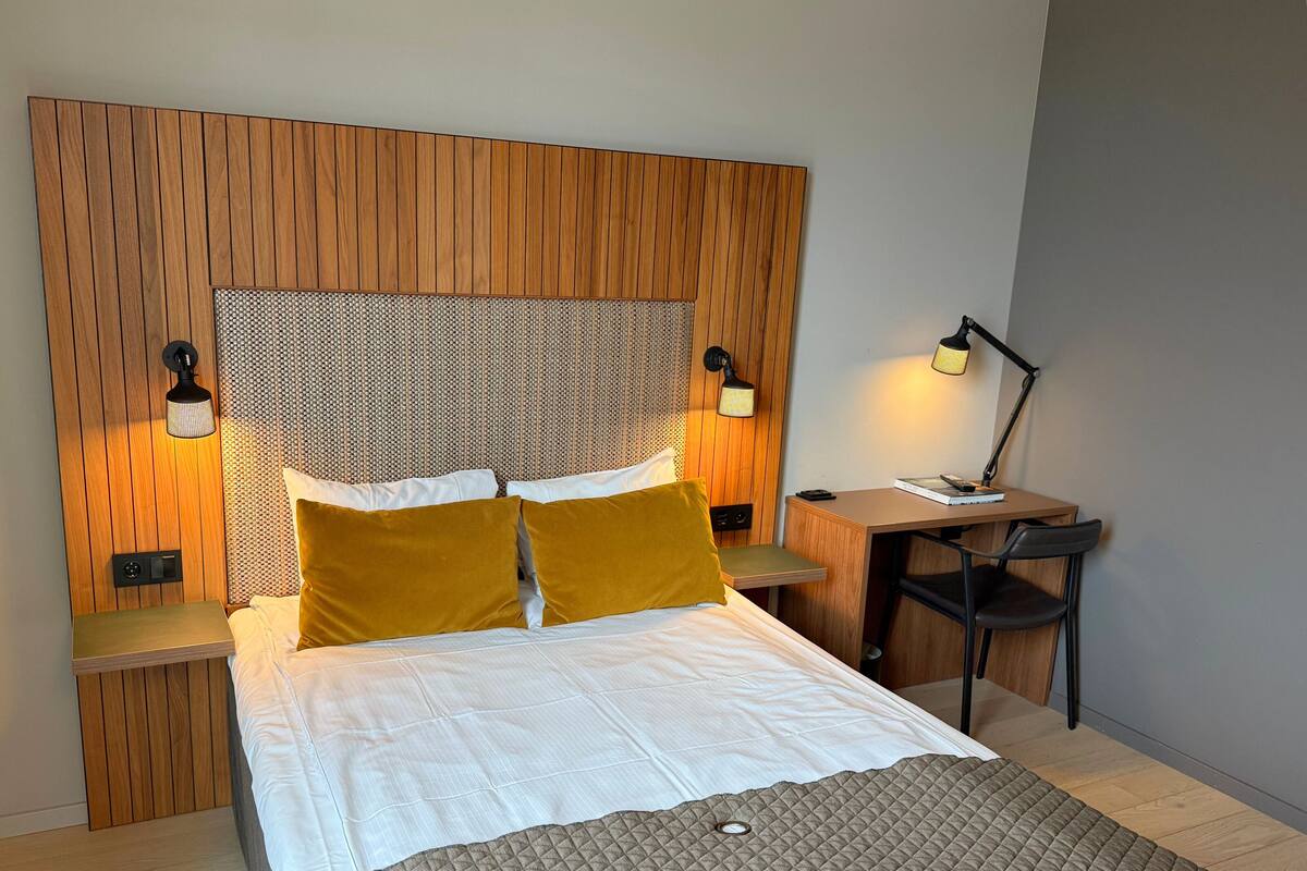 A neatly arranged hotel room with a wooden headboard, a bed with white bedding and mustard pillows, a bedside table with a lamp, and a small desk with a chair.