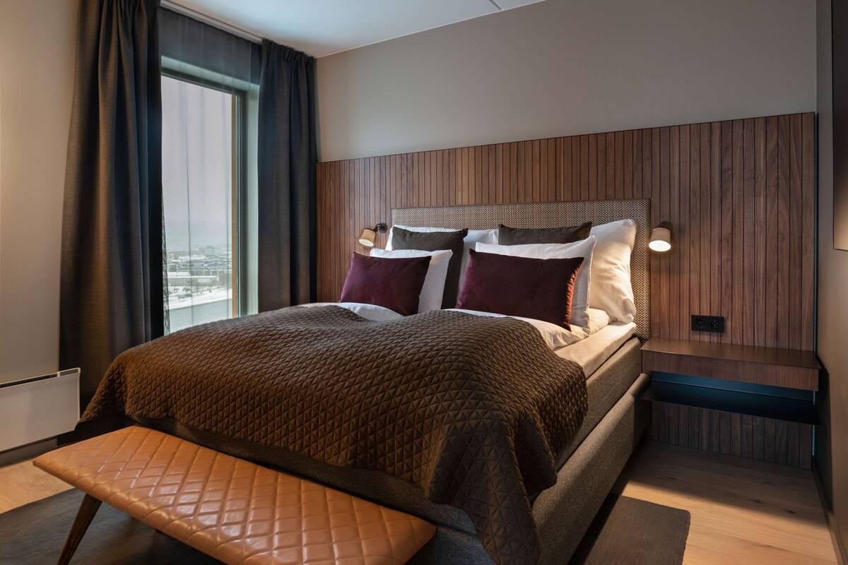 A modern hotel room with a large bed, a bench at the foot, a nightstand with a lamp, and a city view through the window.