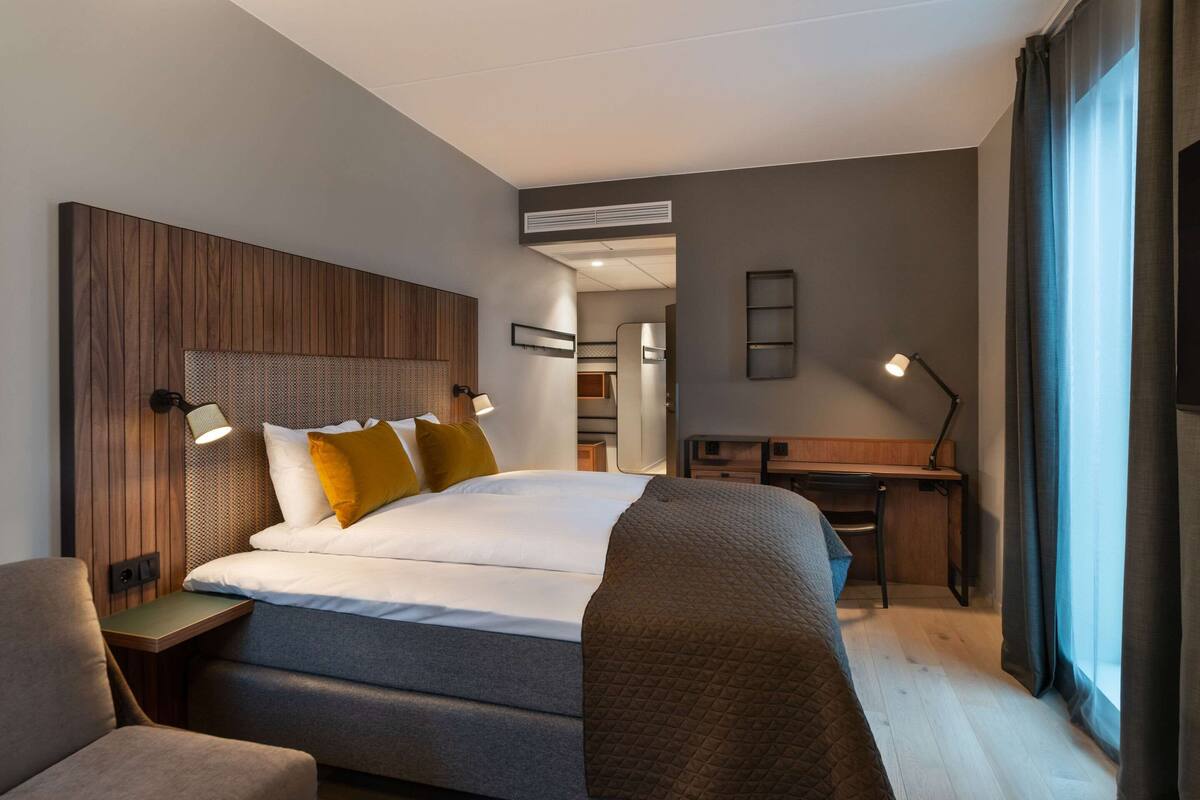 A modern hotel room with a large bed, a desk, and a chair. There is a window with curtains, a wall-mounted lamp, and a ceiling vent.