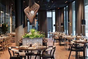 A modern restaurant interior with round tables, dark chairs, and hanging pendant lights. - Quality Hotel River Station (Drammen)