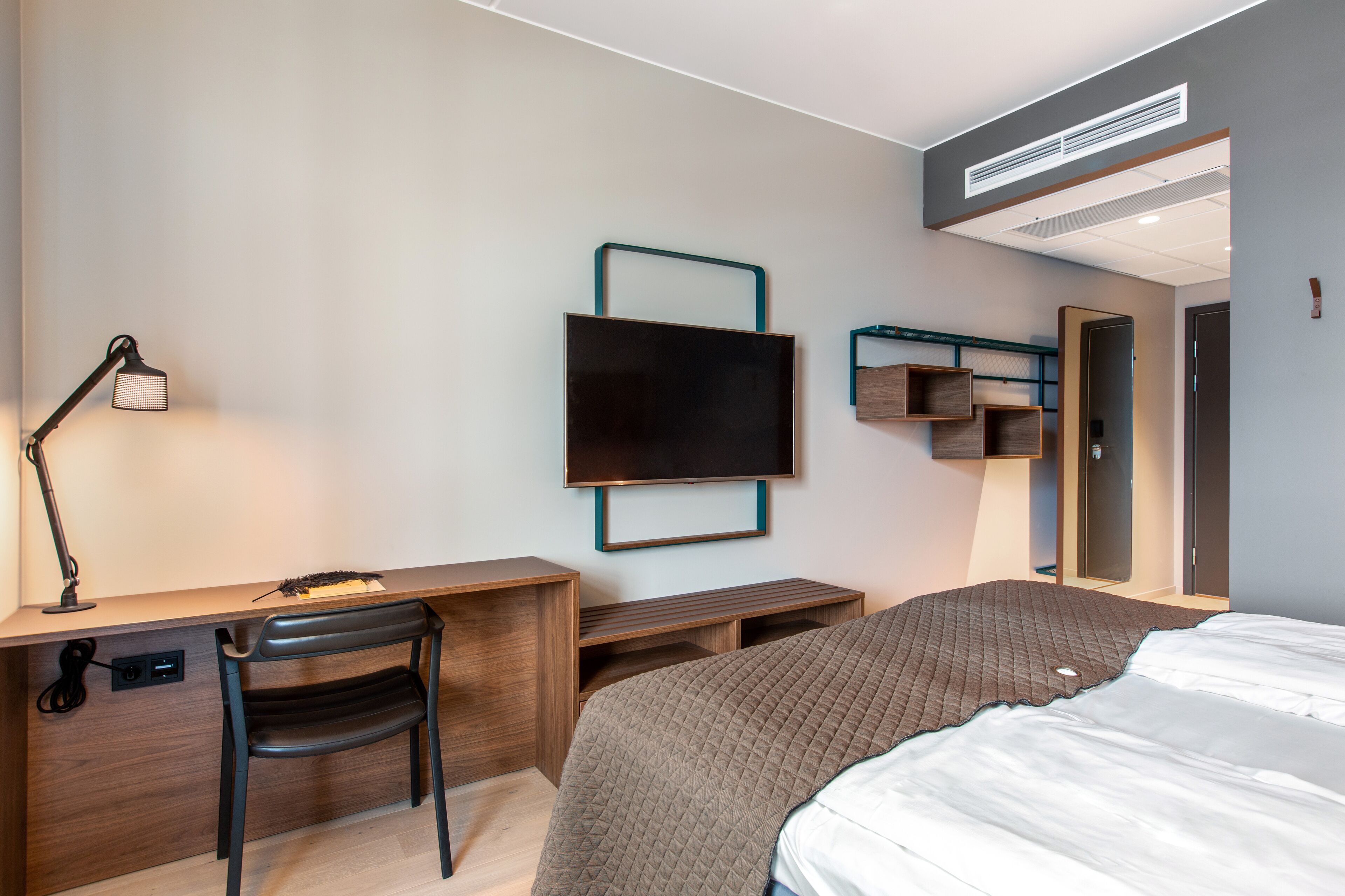 A modern hotel room with a bed, desk, chair, TV, and built-in shelving.