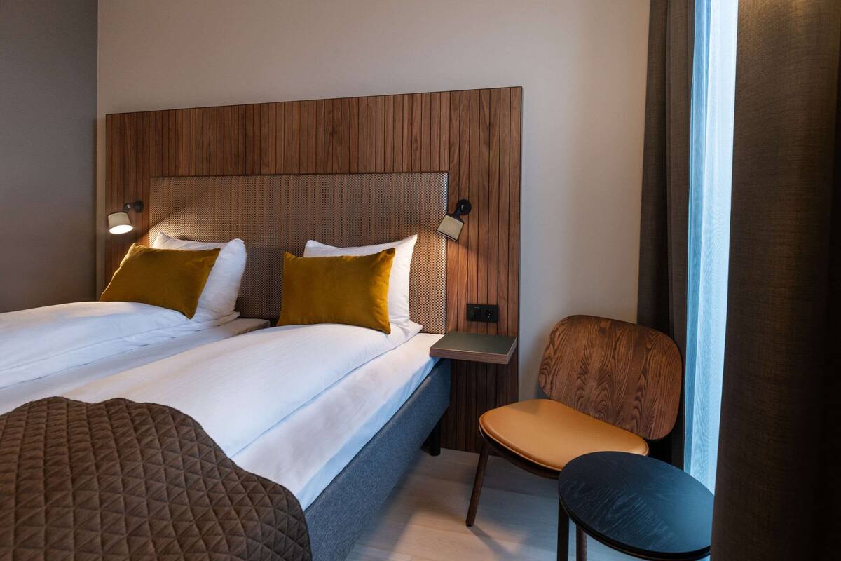 A neatly made bed with two pillows, a bedside table with a lamp, a chair, and a wooden headboard.