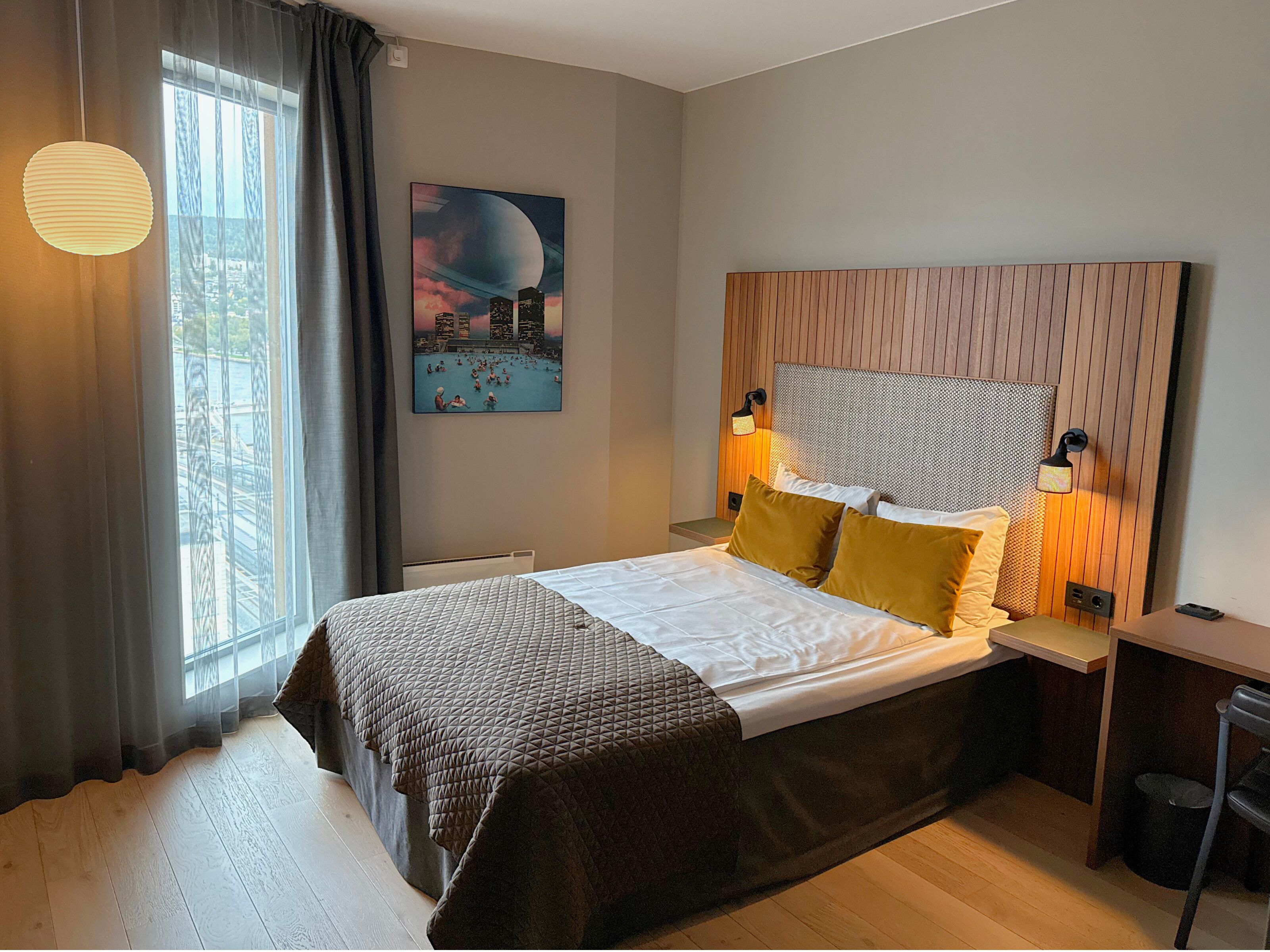 Una habitación de hotel moderna con una cama grande, un cuadro de paisaje urbano, un cabecero de madera y vistas de la ciudad a través de la ventana.