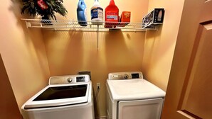 Un cuarto de lavandería con dos lavadoras de carga frontal y un estante superior que contiene diversos productos de limpieza.