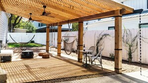 Una pérgola de madera con techo a cuadros, una mesa con sillas y una hamaca.