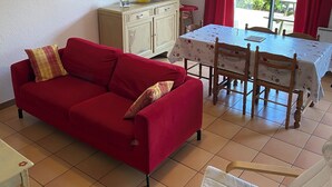 Una sala de estar con un sofá rojo, una mesa de comedor de madera con sillas, un armario y una ventana con cortinas rojas.