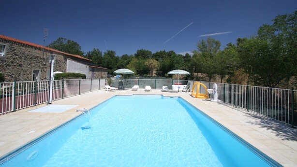 Una piscina rectangular con solárium empedrado, rodeada por una valla de metal blanco. Hay sillones de descanso y sombrillas a lo largo de la piscina.