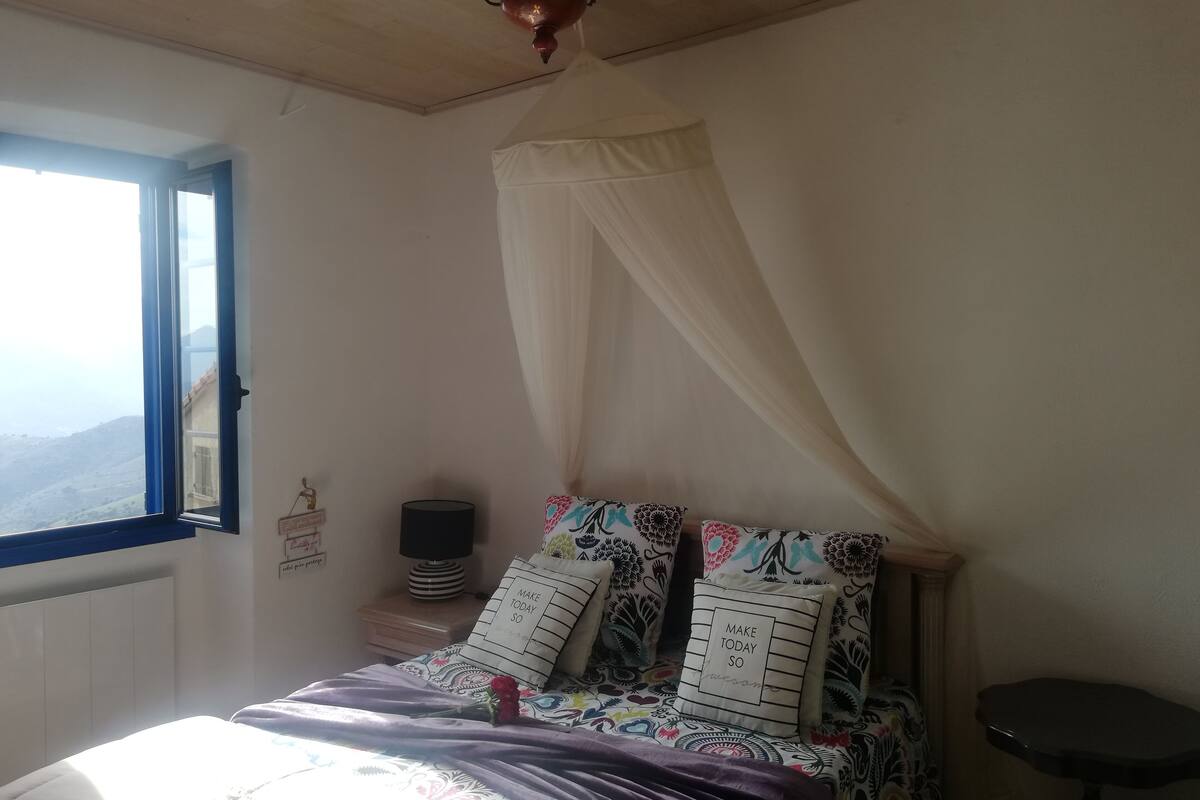 Een slaapkamer met een bed, nachtkastje, lamp en een raam met uitzicht op de omgeving.