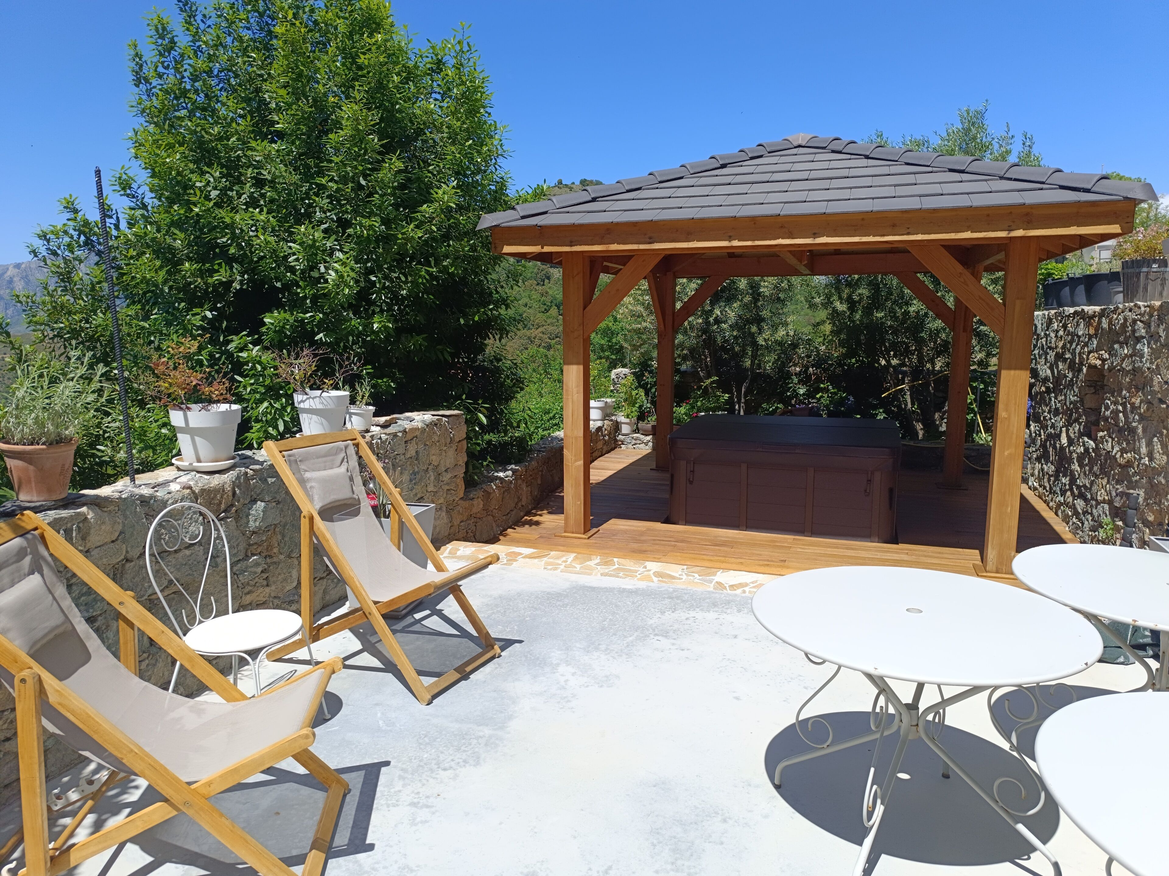 A wooden gazebo with a tiled roof, surrounded by stone walls and potted plants.