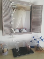 Un dormitorio con un espejo grande, una cama con almohadas blancas y estampadas, una mesita de noche con objetos decorativos y una ventana con cortina.