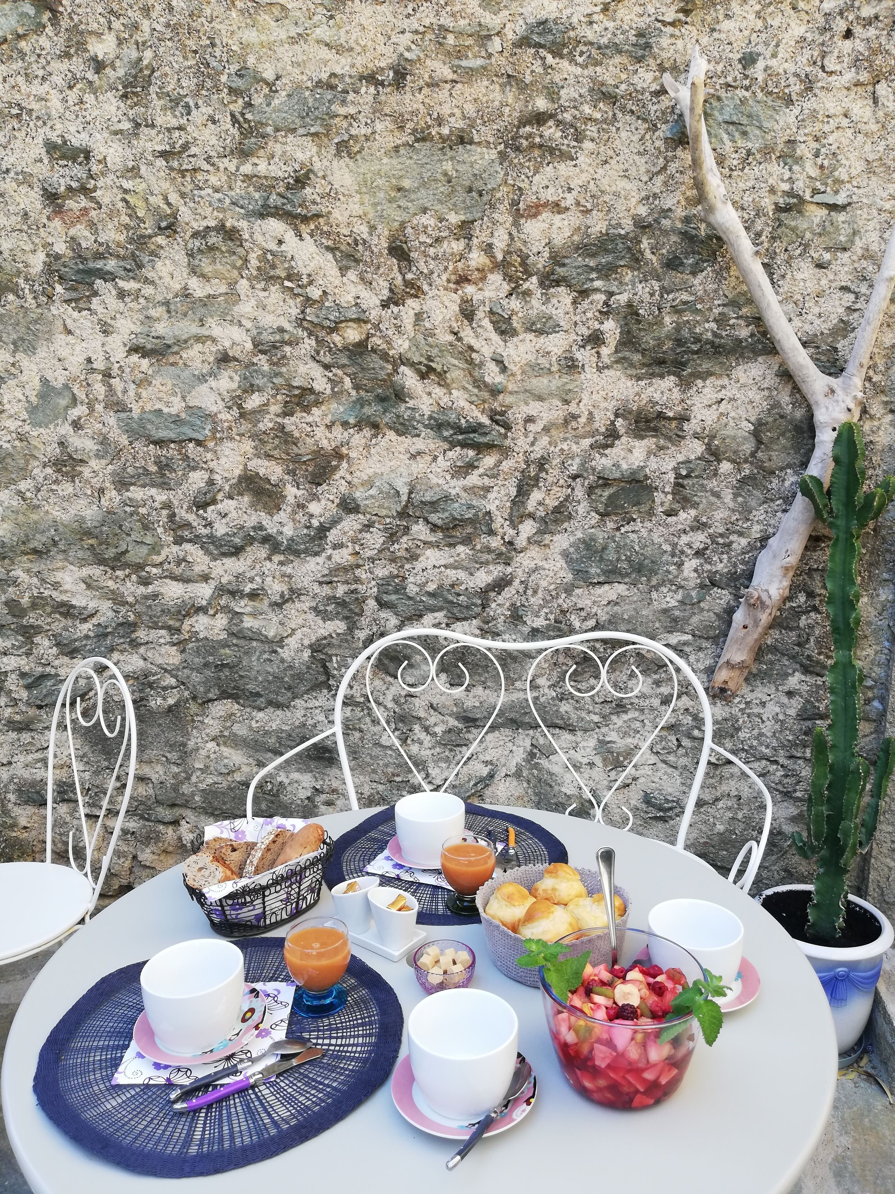 A table set with a variety of food items and drinks, including bread, pastries, and a fruit salad, against a stone wall backdrop.