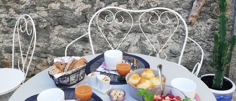 A table set with a variety of food items and drinks, including bread, pastries, and a fruit salad, against a stone wall backdrop.