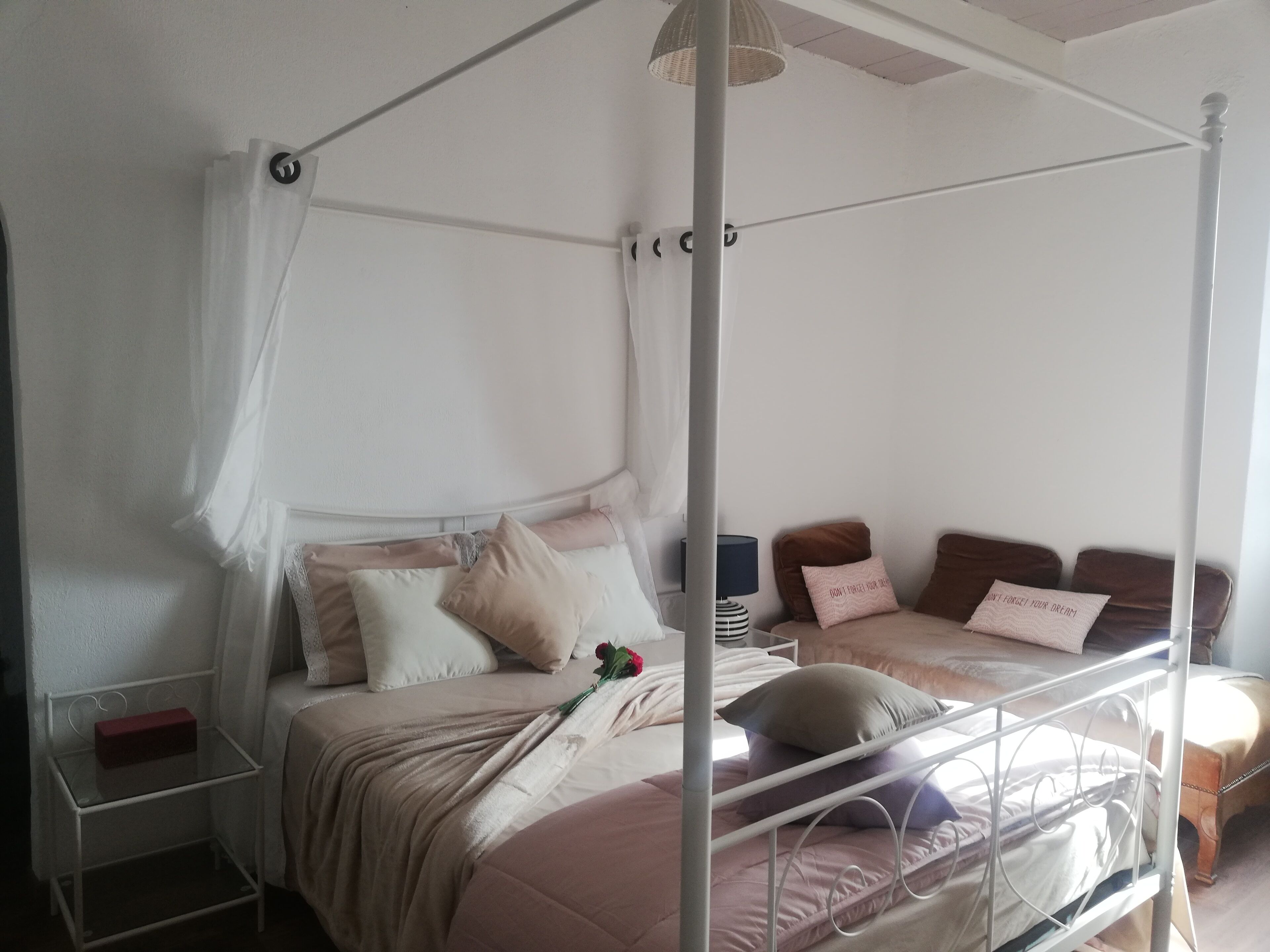 Ein Himmelsbett mit weißem Metallgestell, ein Nachttisch mit Lampe und ein kleiner Nachttisch mit einer roten Schachtel.