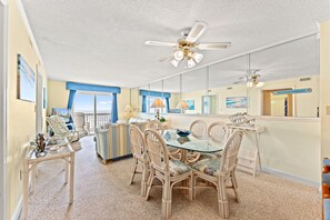 A spacious living area with a glass-top dining table surrounded by wicker chairs, a ceiling fan, and a view of the beach through large windows.