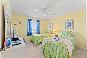 A bedroom with two beds, a ceiling fan, a window with curtains, a desk with a lamp, and a fish-shaped decoration.