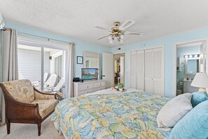 A bedroom with a bed, a chair, a dresser, a ceiling fan, and a bathroom visible through an open door. - Beachmaster 304 Lovely 2 Bedroom Condo With Oceanfront Balcony (NORTH MYRTLE BEACH)