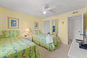 Une chambre d’hôtel avec deux lits, un ventilateur de plafond, des œuvres d’art aux murs et un bureau équipé d’un ordinateur et de télécommandes.