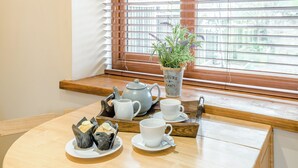 Una mesa de madera con una tetera, tazas y una planta en maceta cerca de una ventana con persianas.