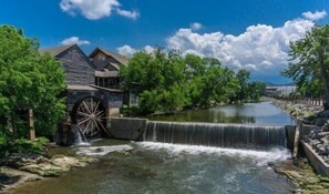 Un molino hidráulico con una rueda grande, rodeado de vegetación exuberante y un río tranquilo.