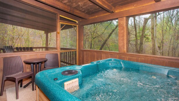 Ein Whirlpool mit Holzterrasse, zwei Korbstühle und Blick auf Bäume durch große Fenster.