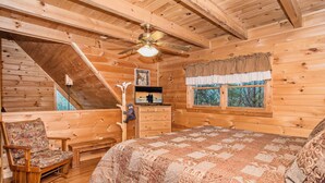 Ein gemütliches Schlafzimmer in einer Blockhütte, mit Holzbalkendecke, einem Bett mit gemustertem Quilt, einem Holzstuhl, einer Kommode und einem Fenster mit Blick auf Bäume.