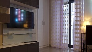 A modern living room with a flat-screen TV mounted on a cabinet, a dining area with a table and chairs, and a large window with sheer curtains. - Casa Nocelle Suite, Boutique and Luxury tiny apartment in the heart of Naples (Napoli)