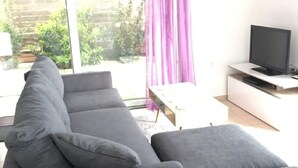 Ein modernes Wohnzimmer mit einer grauen Eckcouch, einem Flachbildfernseher auf einem weißen Lowboard und einem großen Fenster mit violetten Vorhängen.