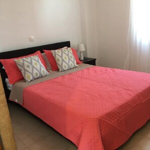 Ein ordentlich bezogenes Bett mit roter Tagesdecke und gemusterten Kissen, eine Nachttischlampe und eine schlichte Wand mit Lichtschalter.