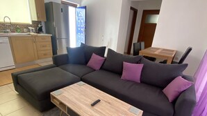 Ein modernes Wohnzimmer mit einem Ecksofa, einem Holztisch und einem im Hintergrund sichtbaren Küchenbereich.