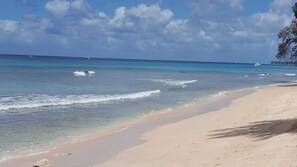 Una playa con agua turquesa cristalina, una orilla arenosa y algunos barcos a lo lejos.