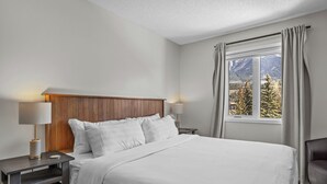 Habitación de hotel con una cama grande, cabecero de madera, dos mesitas de noche con lámparas, vistas a las montañas a través de una ventana y una silla.
