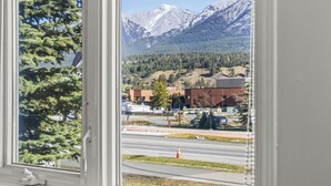Vista a través de una ventana que muestra una calle con edificios, un cono de tráfico y montañas nevadas al fondo.