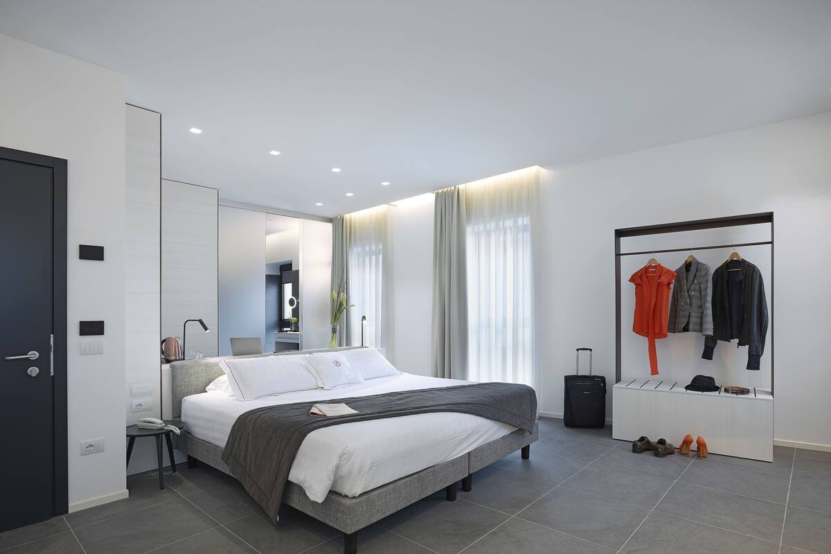 Ein modernes Hotelzimmer mit einem großen Bett, einem Kleiderschrank, einem Schminktisch mit Spiegel und einem Kleiderständer mit drei Jacken.