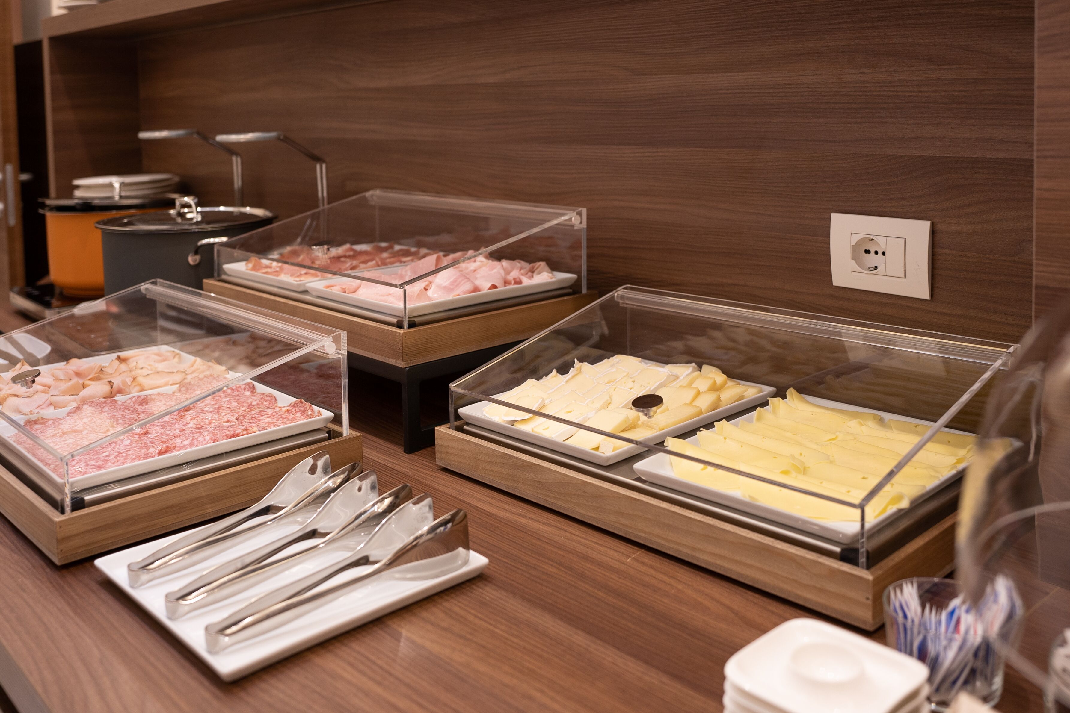 A buffet with various food items in clear containers, including meats and cheeses, with utensils laid out on a table.